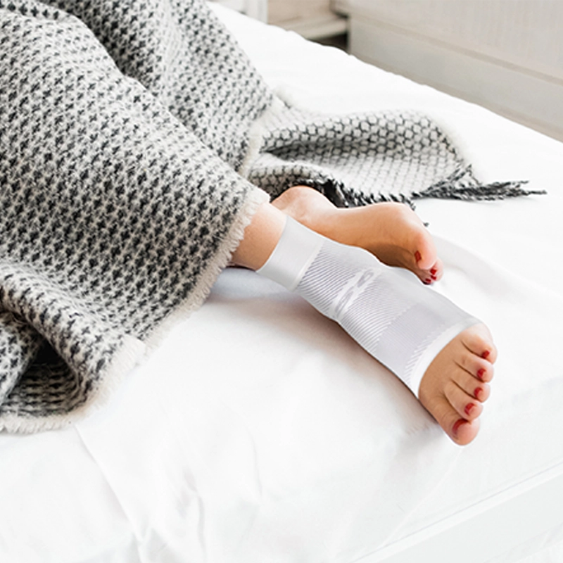 Close up of woman's feet poking out of a blanket with the nigh time Plantar Fasciitis sleeve on
