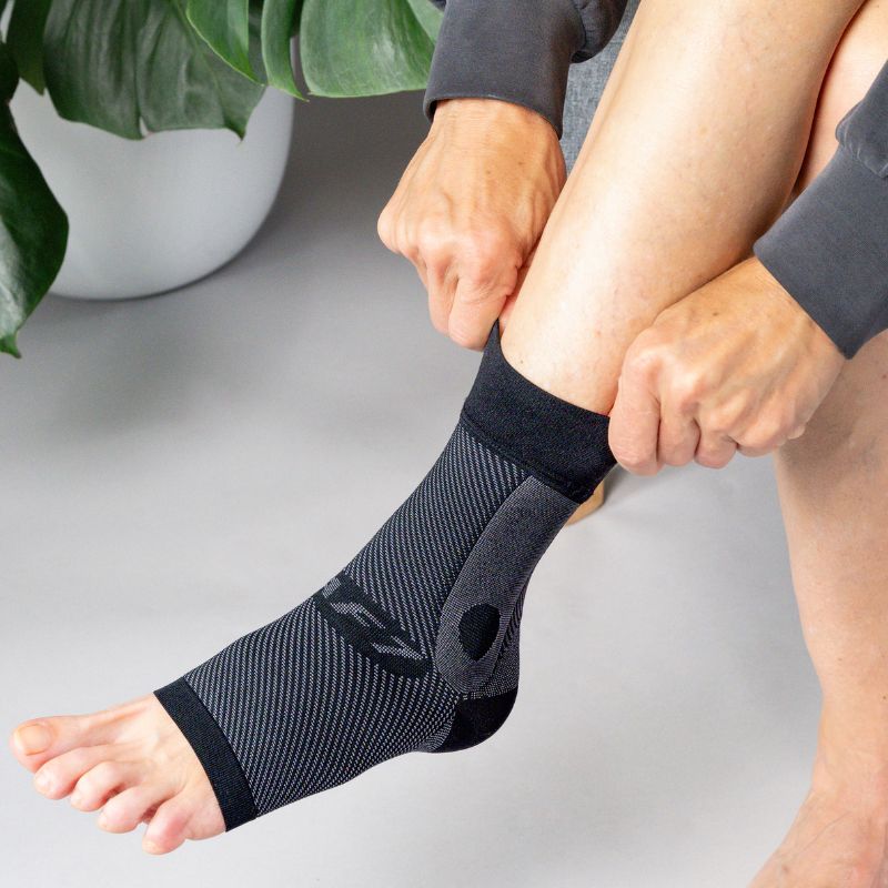 Person applying a black ankle sleeve on a light gray surface with a plant in the background.