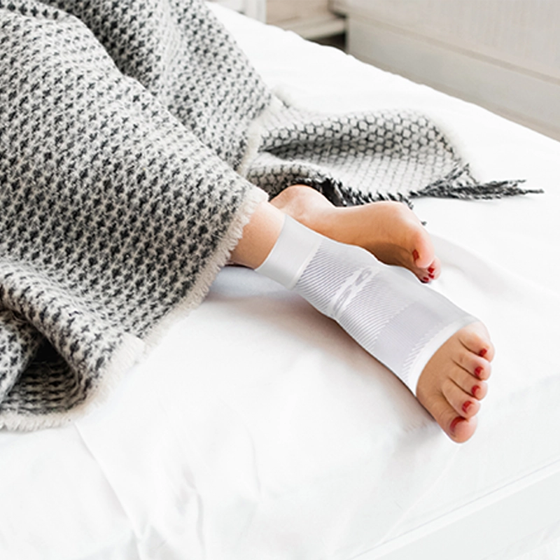Close up of woman's feet poking out of a blanket with the night time Plantar Fasciitis sleeve on