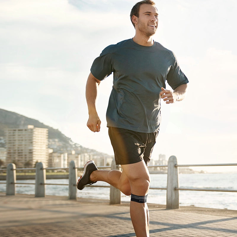 Image of a man running on a side walk next to the ocean while wearing the patella compression bracing sleeve
